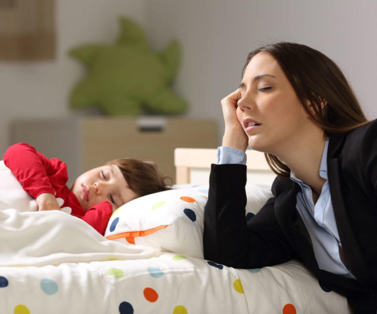 Tired worker mother wearing suit sleeping beside her sleepy daughter on a bed at home